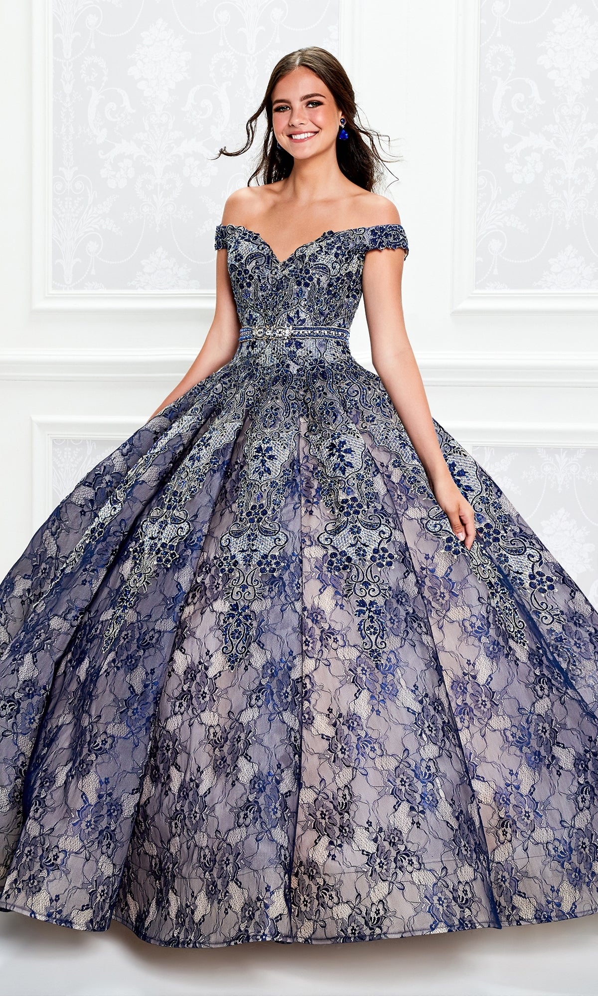 A young woman smiles against a white ornate background wearing the Princesa Lace Ball Gown Quinceañera Dress PR11921, an off-the-shoulder, floor-length gown with intricate blue and silver lace detailing.